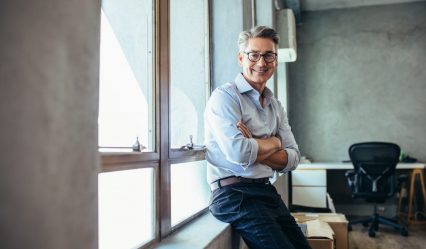 Smiling businessman looking at camera as he stands by a window sill with arms crossed. Mid adult businessman at his office.