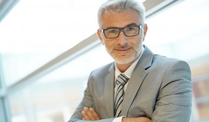 Stylish mature businessman leaning on office window looking at camera